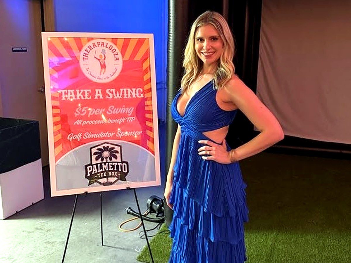Host beside a Take a Swing sponsor sign at a fundraiser with the Palmetto Tee Box bay behind