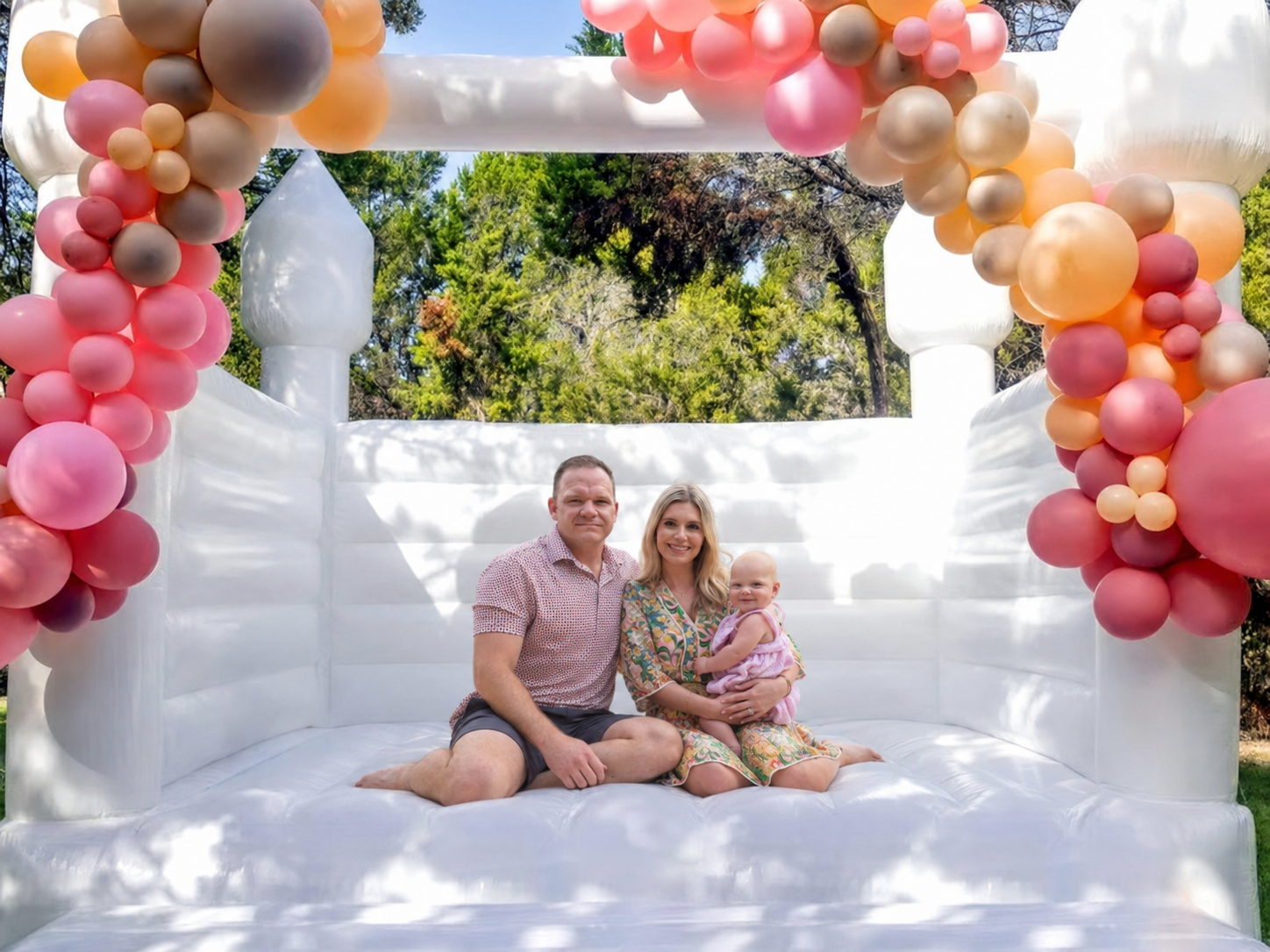 A family of three sitting inside a white Hartwell Inflatables bounce house beneath a blush and peach balloon arch